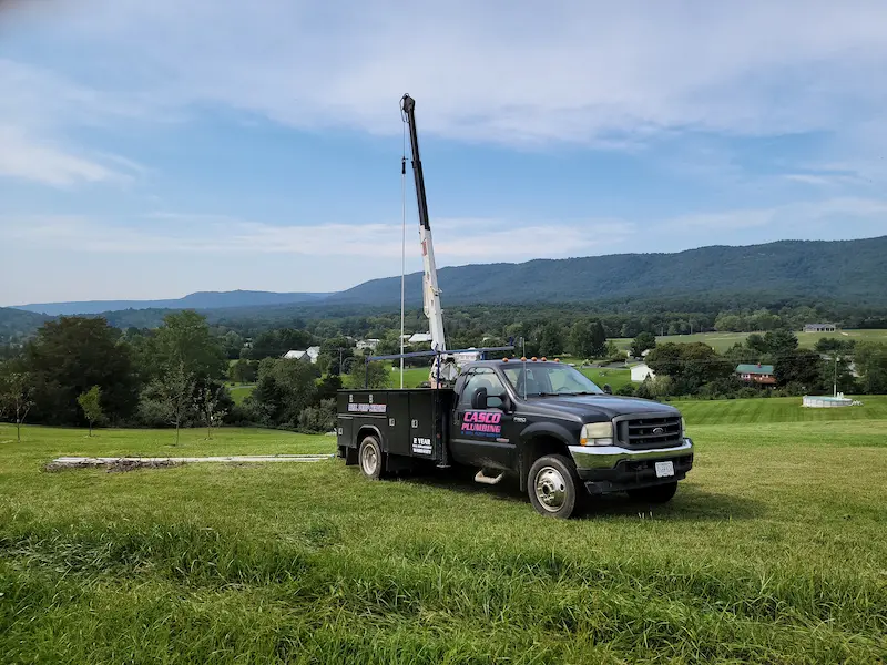 Casco Plumbing truck with crane arm on a green field, mountains in background.